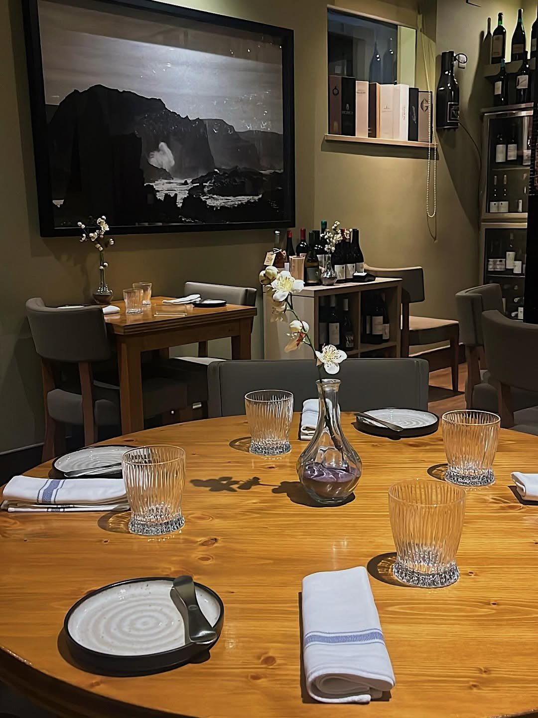 Mesa redonda de madeira posta com copos, guardanapos e uma jarra com flores, ao lado de prateleiras com garrafas de vinho