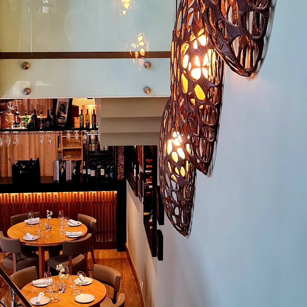 Sala vista de cima, com mesas redondas postas, balcão iluminado e candeeiros de parede em forma de folha